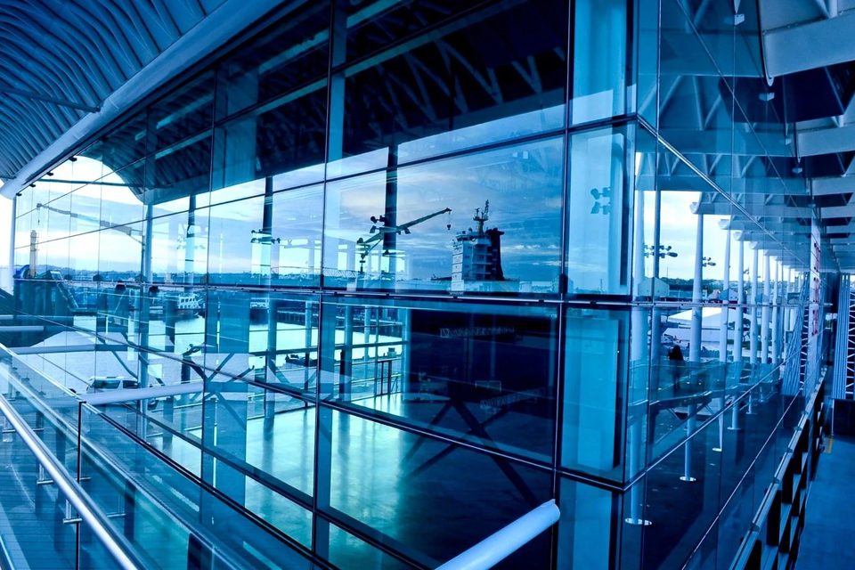 Fachada de cristal de una terminal portuaria, con el muelle y una grúa reflejados en los paneles