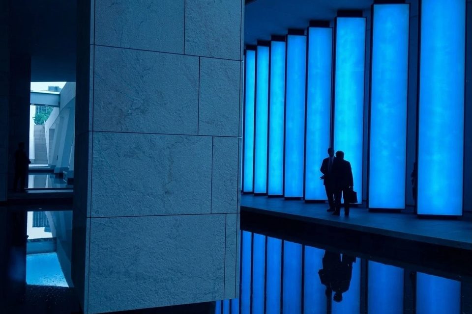 Vestíbulo en penumbra con una hilera de columnas de luz azul reflejadas en el suelo pulido, y dos personas frente a ellas